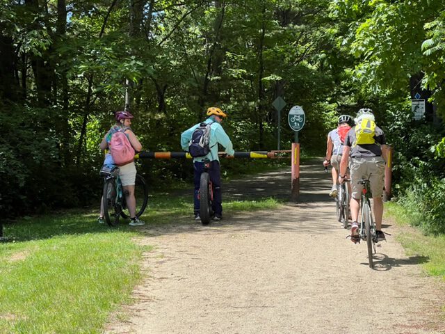 cyclists ride along the Cheshire Rail Trail North, a path that is packed dirt with many trees